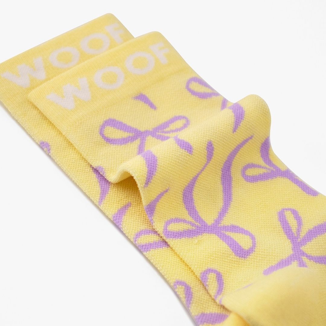 Macro close-up shot showing the Bows Bamboo Crew Compression Socks folded neatly to highlight the white woven "WOOF" cuff bands and the purple ribbon bow knit pattern layout.
